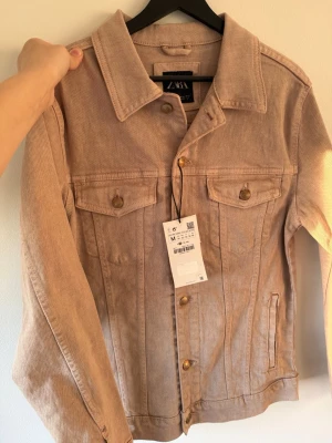 Beige jeansjacka från Zara - Snygg beige jeansjacka från Zara med klassisk krage, två bröstfickor med lock och knappar i guldton. Jackan har raka ärmar och stilrena sömmar som ger en clean look. Perfekt att slänga över en hoodie eller t-shirt för en avslappnad vibe.