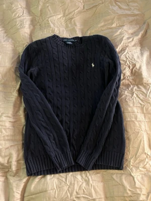 Mörkblå kabelstickad tröja Polo Ralph Lauren - Kabelstickad mörkblå tröja från Polo Ralph Lauren med klassisk gul broderad logga på bröstet. Tröjan har rund halsringning, långa ärmar och ribbade muddar. Perfekt för dig som gillar tidlös och stilren design. Tröjan har en slitning i kragen