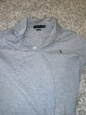 Grå pikétröja från Polo Ralph Lauren - Snygg grå pikétröja från Polo Ralph Lauren i slim fit-modell. Klassisk krage, knappslå och den ikoniska broderade loggan på bröstet. Perfekt till jeans eller chinos för en clean och stilren look.