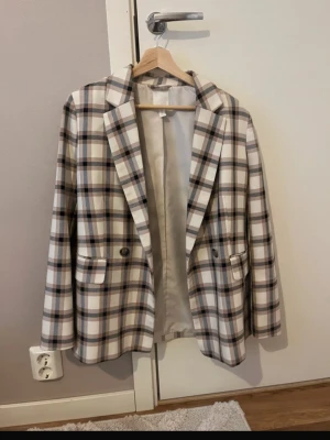 Kavaj - Snygg kavaj med rutigt mönster i beige, brunt och vitt. Klassisk passform med två knappar framtill och fickor med lock. Fodrad insida för extra komfort. Perfekt för dig som vill ha en trendig och stilren look.