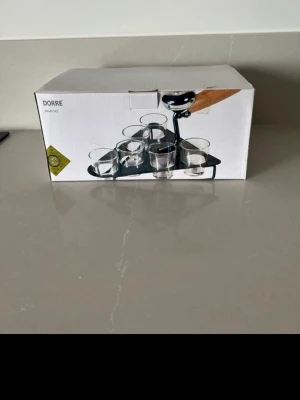 Dorre snaps set med glas och ställ - Säljer ett snaps set från Dorre som innehåller flera små glas och ett svart ställ. Perfekt för att servera shots eller snaps på ett snyggt sätt. Glasen är genomskinliga och stället har en modern design, utformad som en murslev med handtag i trä.