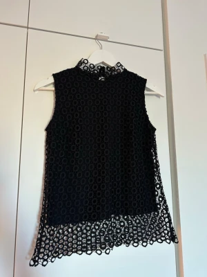 Svart spetsblus med hög krage - Säljer en svart ärmlös blus med hög krage och vackert spetsmönster. Blusen har en liten öppning bak i nacken. Perfekt för dig som gillar detaljer och en elegant look.