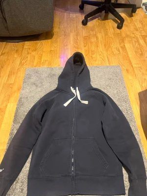 Mörkblå hoodie med dragkedja från russemerch - Mörkblå hoodie från Russe merch med vit dragsko och dragkedja framtill. Klassisk modell med kängurufickor och ribbade muddar vid ärmslut och nederkant. Tillverkad i mjukt bomullsmaterial, perfekt för en avslappnad stil.