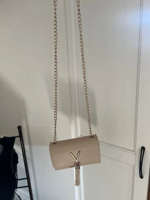 Beige axelväska från Valentino - Snygg beige axelväska från Valentino med guldfärgad kedjerem och dekorativt V-spänne framtill. Väskan har en elegant tofsdetalj och är tillverkad i ett strukturerat konstläder. Perfekt för dig som vill ha en stilren och trendig accessoar.