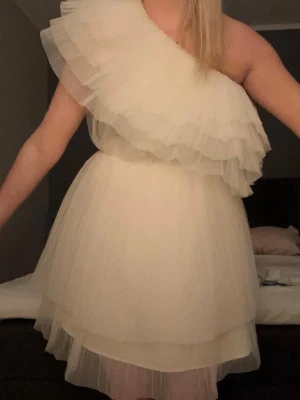 Vit oneshoulder volangklänning - Supersöt vit klänning med lager på lager av volanger och oneshoulder-design. Klänningen har en fluffig, luftig känsla och är perfekt för dig som vill sticka ut. Tillverkad i ett tunt, lätt material som ger en romantisk vibe.