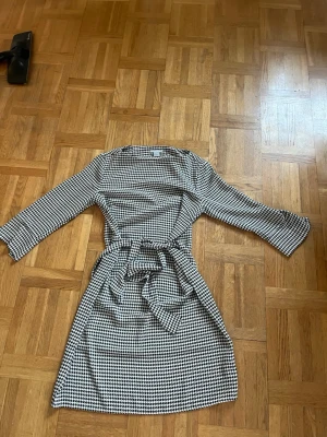 Rutig kort klänning med knytband - Snygg svartvit rutig klänning med trekvartsärm och knytband i midjan. Klänningen har en klassisk, rak passform och är tillverkad i ett mjukt material som känns bekvämt mot huden. Perfekt för dig som gillar stilrena mönster och enkel design.