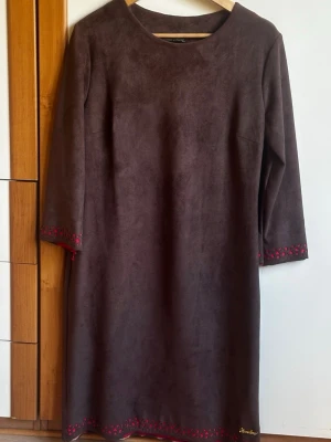 Brun mockaklänning från Kenn More - Brun långklänning från Kenn More i mjuk mocka-look. Klänningen har trekvartsärm och dekorativa röda broderier vid ärmslut och nederkant. Enkel, rak passform och rund halsringning. Perfekt för dig som gillar stilrena detaljer.