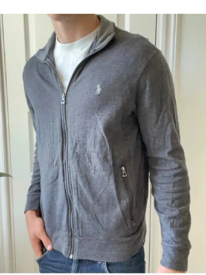 Grå zip-tröja från Polo Ralph Lauren - Snygg grå zip-tröja från Polo Ralph Lauren med diskret logga på bröstet. Tröjan har hel dragkedja, två fickor med zip och ribbade muddar. Perfekt att slänga på över en t-shirt för en avslappnad look.