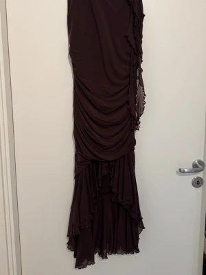 Brun långklänning med volang - Elegant brun långklänning med draperad design och volangdetaljer längs sidan och nederkanten. Klänningen har ett asymmetriskt fall och är tillverkad i ett lätt, flödande material som ger ett snyggt och dramatiskt intryck. Perfekt för dig som vill sticka ut.