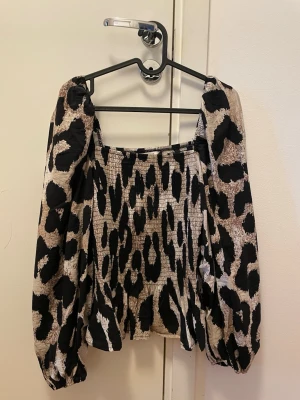 Leopardmönstrad topp med puffärmar - Trendig topp med leopardmönster i svart och beige. Modellen har puffiga långärmar och smockad, åtsittande kropp. Fyrkantig ringning och ballongärmar ger en cool och modern look. Perfekt för dig som vill sticka ut.