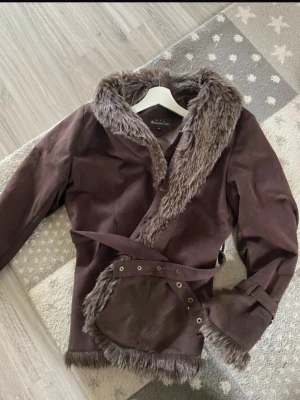 Brun pälsjacka med bälte - Säljer en brun pälsjacka från Gina Tricot med mjuk fuskpäls längs krage, ärmslut och nederkant. Jackan har ett matchande bälte i midjan och dekorativa knappar. Perfekt för kalla dagar och ger en snygg vintagekänsla. Storlek 40 så M-L