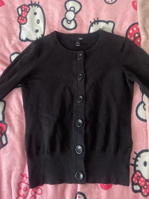 Svart kofta från H&M med knappar - Klassisk svart kofta från H&M med rund halsringning och fem stora svarta knappar framtill. Koftan har ribbade muddar vid ärmslut och nederkant. Tillverkad i mjukt och stretchigt material som passar perfekt till lager på lager.