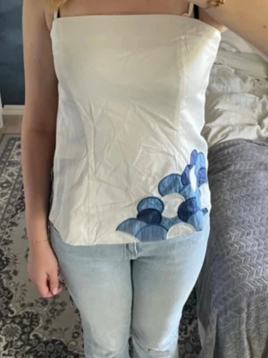 Vit korsettopp med blå detaljer - Säljer en vit korsettopp med raka linjer och tunna axelband. Toppen har en unik design med blå och ljusblå cirkelformade detaljer på ena sidan. 