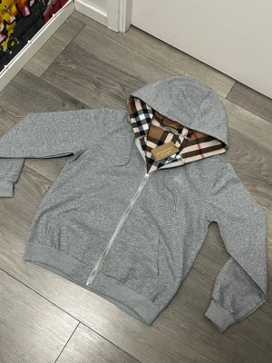 Grå zip hoodie från Burberry - Snygg grå hoodie med dragkedja från Burberry. Klassisk rutmönstrad huva i beige, svart, vitt och rött. Broderad logga på bröstet och ribbade muddar vid ärmar och midja. Perfekt för en stilren och avslappnad look.