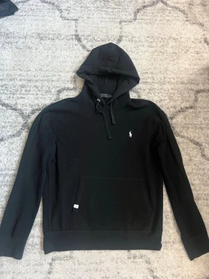 Svart hoodie från Polo Ralph Lauren - Klassisk svart hoodie från Polo Ralph Lauren med huva och dragsko. Broderad vit logga på bröstet och stor magficka framtill. Tillverkad i mjukt bomullsmaterial som känns skönt mot huden. Perfekt för en avslappnad och stilren look.