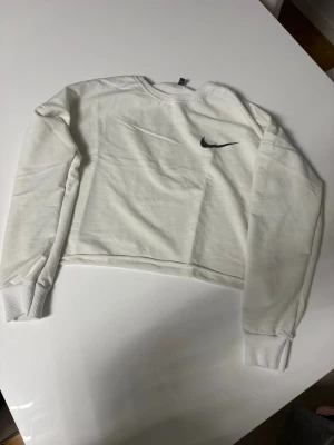 Vit cropped sweatshirt Nike - Säljer en vit cropped sweatshirt från Nike med svart Swoosh-logga på bröstet. Tröjan har långa ärmar, ribbade muddar och rund halsringning. Perfekt för en sportig och avslappnad stil. Materialet känns mjukt och bekvämt.  Not real nike
