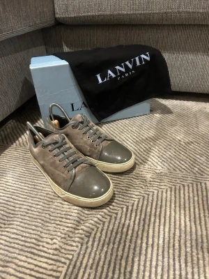 Gråa Lanvin sneakers i mocka och lack - Snygga sneakers från Lanvin i grå mocka med matchande grått lack på tån och häl. Skorna har en beige sula och grå skosnören. Klassisk låg modell med stilren design och exklusiv känsla. Perfekta för dig som gillar lyxiga detaljer och tidlös stil. Box och påse ingår ej 