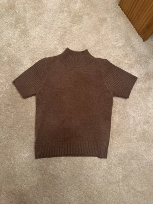 Brun stickad tröja med kort ärm - Snygg brun stickad tröja med korta ärmar och hög ribbad krage. Tröjan har ribbade detaljer vid ärmslut och nederkant, vilket ger en stilren look. Perfekt att matcha med jeans eller kjol för en trendig outfit. Aldrig använd!!!