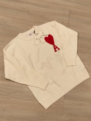 Vit stickad tröja från Ami Paris - Vit stickad tröja från Ami Paris med röd hjärt- och A-detalj på bröstet. Tröjan har rund halsringning, ribbade muddar och nederkant. Perfekt för dig som gillar stilrena plagg med en twist.