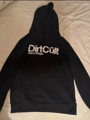 Svart Dirt Cult hoodie med tryck - Svart hoodie från Dirt Cult med stor vit logga och texten 'San Diego' på bröstet. Klassisk känguruficka framtill och justerbar huva med dragsko. Perfekt för dig som gillar streetwear och vill ha en stilren look.