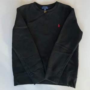 Svart sweatshirt från Polo Ralph Lauren - Klassisk svart sweatshirt från Polo Ralph Lauren med rund halsringning och den ikoniska röda logotypen broderad på bröstet. Tröjan har långa ärmar och ribbade muddar vid ärmslut och nederkant. Perfekt för en avslappnad och stilren look.