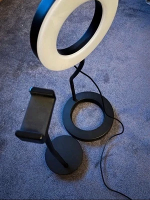 Ring light med mobilhållare USB - Säljer en svart ring light med mobilhållare och USB-anslutning. Lampan har rund form och justerbar arm, perfekt för streaming, videos eller selfies. Styrs med fjärrkontroll på sladden för olika ljuslägen. Passar de flesta smartphones.