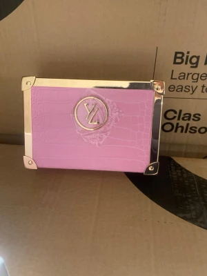 Rosa kuvertväska med gulddetaljer - Snygg rosa kuvertväska i boxformad modell med krokopräglat mönster och guldfärgade metalldetaljer. Väskan har ett stort LY-emblem framtill och förstärkta hörn i metall. Insidan är ljus och har plats för småsaker. Perfekt accessoar för att lyfta din outfit.//  byta batteri