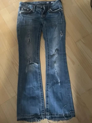 Blå bootcut jeans med slitningar - Säljer ett par blå bootcut jeans från Miss Me med slitna detaljer och dekorativa sömmar. Jeansen har låg midja, broderade bakfickor och råa kanter vid bensluten. Materialet är denim i bomull med stretch för skön passform. Bildena är ifrån tidigare ägare❤️ säljer då de va för små 💕