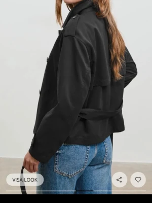 Kort svart trenchcoat med bälte - Stilren svart trenchcoat i kort modell med klassisk krage, axelklaffar och knytskärp i midjan. Jackan har dubbelknäppning och detaljer som ger en tidlös look. Perfekt att slänga över en hoodie eller t-shirt för en cool vibe.