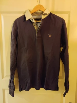 Mörkblå rugby piké från GANT storlek M - Säljer en mörkblå henley från GANT med vit kontrastkrage och knappslå. Skjortan har lång ärm och diskret GANT-logga broderad på bröstet. Perfekt för dig som gillar klassisk och stilren design. Storlek M