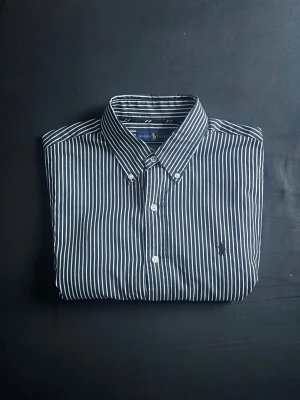 Svart/Vit skjorta från Ralph Lauren - Snygg svart och vit randig skjorta från Ralph Lauren i slim fit. Skjortan är tillverkad i 100% bomull med stretch och har klassisk krage samt knappar framtill. Perfekt nu inför sommaren. Tveka inte med frågor eller prisförlsag