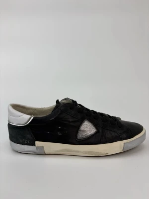 Philippe Model svarta sneakers - Svarta sneakers från Philippe Model med vit sula, vit hälkappa och silverfärgad detalj på sidan. Skorna har klassisk låg profil, svarta skosnören och är tillverkade i läder. Perfekta för dig som gillar en stilren men ändå unik look.