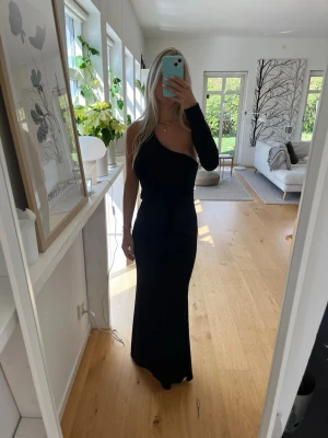 Svart långklänning oneshoulder - Svart långklänning med oneshoulder-design och lång ärm på ena sidan. Klänningen har en figurnära passform och är perfekt för dig som vill sticka ut med en stilren och modern look. Materialet känns mjukt och följsamt mot kroppen. Du kan välja att dra ärmen runt halsen istället så det ser mer ut som halterneck😊