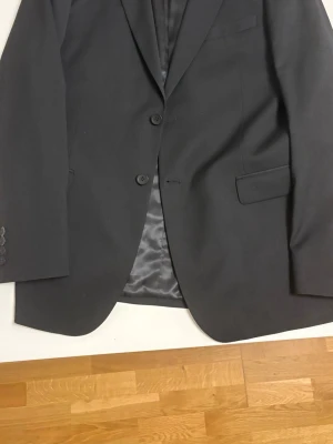Svart kostym med tvåknäppt kavaj - Stilren svart kostym med klassisk tvåknäppt kavaj och matchande byxor. Kavajen har fickor med lock och slät finish. Perfekt för dig som vill ha en tidlös look. Materialet känns mjukt och följsamt. Använd endast en gång säljer för har inget behov. Storlek är 54 på kavajen och på byxor  38-32.