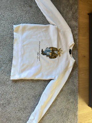 Vit Polo Bear hoodie från Ralph Lauren - Vit hoodie från Ralph Lauren med ikoniska Polo Bear-trycket på bröstet. Björnen har cowboyhatt, brun fransjacka och jeans. Klassisk passform med långa ärmar och rund hals. Perfekt för dig som gillar streetstyle och snygga prints.