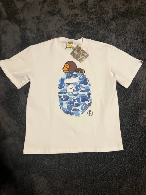 Vit BAPE t-shirt med blått camo-tryck - Vit bape tröja med blått camo tryck på framsidan. Tröjan är helt oanvänd med lapp kvar. Hör av er vid frågor 