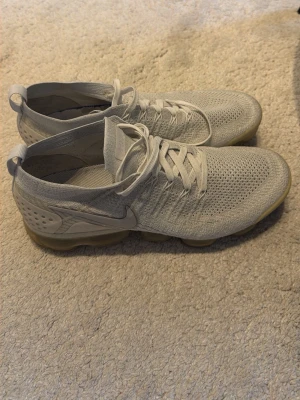 Nike Air Vapormax  vit - Säljer ett par Nike Air Vapormax Flyknit sneakers i ljusgrått. Skorna har en stickad ovandel i mesh för extra andningsförmåga och en genomskinlig sula med synliga luftkuddar. Snygg och modern design med klassisk Nike-logga på sidan.