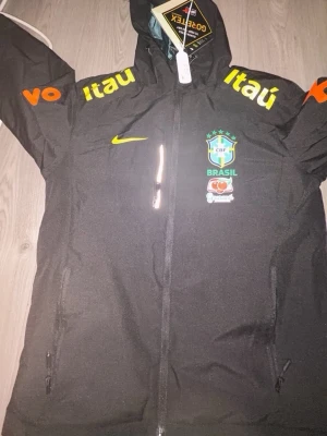 Svart Brasil Nike vindjacka Gore-Tex - Svart vindjacka från Nike med Brasil CBF-logga, gula och orange tryck på ärmar och bröst, samt GORE-TEX-material. Jackan har huva, hel dragkedja och två sidofickor med dragkedja. Perfekt för dig som gillar sportig stil och vill sticka ut.