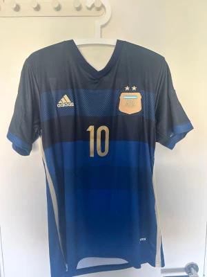 Argentina Messi matchtröja  - Snygg blå Argentina matchtröja från Adidas med Messi och nummer 10 i guld på ryggen. Tröjan har korta ärmar, V-ringning och två stjärnor ovanför landslagsemblemet. Tillverkad i lätt och ventilerande material, perfekt för fotboll eller träning. Lappen kvar och pris går och diskutera