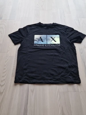 Svart Armani Exchange t-shirt - Svart t-shirt från Armani Exchange med tryck framtill i form av två rutor med bokstäverna 'A' och 'X' samt texten 'Armani Exchange' under. Klassisk passform och rund hals. Perfekt för en stilren och modern look.