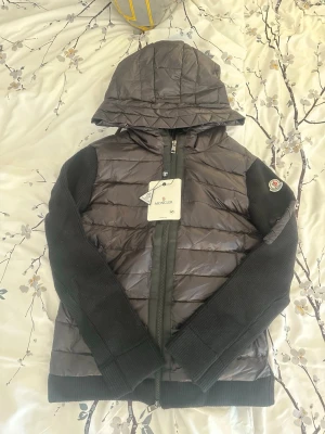 Moncler Cardigan - Snygg och exklusiv Moncler jacka i svart modell med kombination av stickat material och lätt dunframtill. Jackan har huva, dragkedja framtill samt diskret Moncler-logga på ärmen. Väldigt bekväm och perfekt för både vardag och finare outfits.