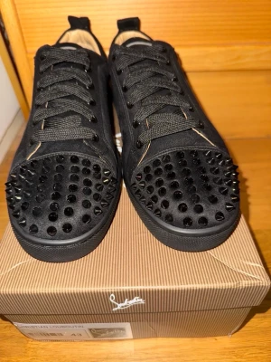 Svarta Louboutin sneakers med nitar - Säljer ett par svarta sneakers från Christian Louboutin med mattsvarta nitar på tån och klassisk röd sula. Skorna har snörning, ovandel i mocka och coola detaljer som sticker ut. Perfekta för dig som vill ha något extra till din outfit.