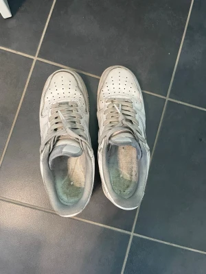 Nike Air Force 1 vita sneakers - Klassiska Nike Air Force 1 sneakers i helvitt läder med grå detaljer. Skorna har perforerad tå, kraftig sula och ikonisk Swoosh-logga på sidorna. Perfekta för en stilren och sportig look.