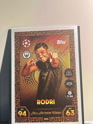 Topps fotbollssamlarkort Rodri All Action Hero 25/26 match attack - Samlarbild från Topps med Rodri i Manchester Citys matchtröja. Kortet har bronsfärgad ram, grafiska detaljer och texten 'All-Action Hero'. Siffror och symboler för försvar och anfall finns på kortet. Rodri All Action Hero 25/26 match attack