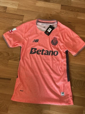 Porto rosa matchtröja New Balance - Säljer en rosa FC Porto matchtröja från New Balance med svarta detaljer och tryck. Tröjan har klubbmärke, sponsorloggor och mönstrad yta. Korta ärmar med randiga muddar och officiella Liga Portugal-märket på ärmen. Perfekt för fotbollsfans!