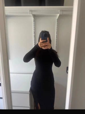 Svart ribbad långklänning - Svart långklänning med ribbad struktur och lång ärm. Klänningen har en tight passform och en slits på sidan som ger en snygg detalj. Perfekt för dig som gillar stilrena och enkla plagg med en modern touch.
