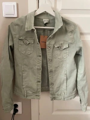 Ljusgrön jeansjacka - Snygg ljusgrön jeansjacka med klassisk krage, två bröstfickor med lock och knapp samt silverfärgade knappar framtill. Jackan har raka ärmar och en avslappnad passform som passar perfekt till många olika stilar. Märket är Onado och jag köpte från Sellpy 