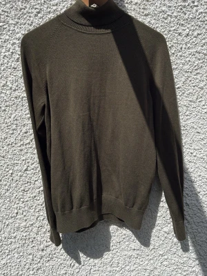 Olivgrön polotröja från Bläck - Snygg olivgrön polotröja från Bläck med ribbade muddar och hög krage. Tröjan är tunnstickad och har en stilren, minimalistisk design som passar perfekt till lager-på-lager. Materialet känns mjukt och följsamt.