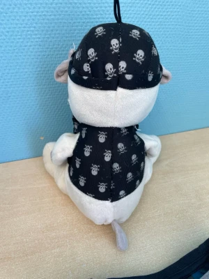Plyschhund med pirattema - Mjuk och gosig plyschhund med grå och vit päls. Hunden har ett svart piratbandana och väst med dödskallemönster samt ett rött hjärtformat ögonlapp. Perfekt som rolig present eller dekoration.