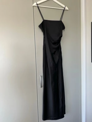 Svart långklänning med smala axelband - Elegant svart långklänning med smala axelband och rak siluett. Klänningen har en lätt glansig finish och är tillverkad i ett slätt material som ger en lyxig känsla. Perfekt för dig som gillar stilrena och minimalistiska plagg.